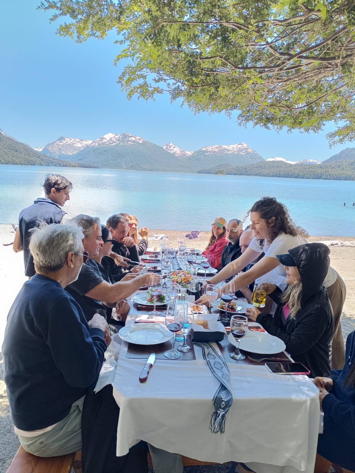 Mesa larga a la orilla del lago bajo un árbol, con un grupo de personas comiendo y montañas nevadas de fondo