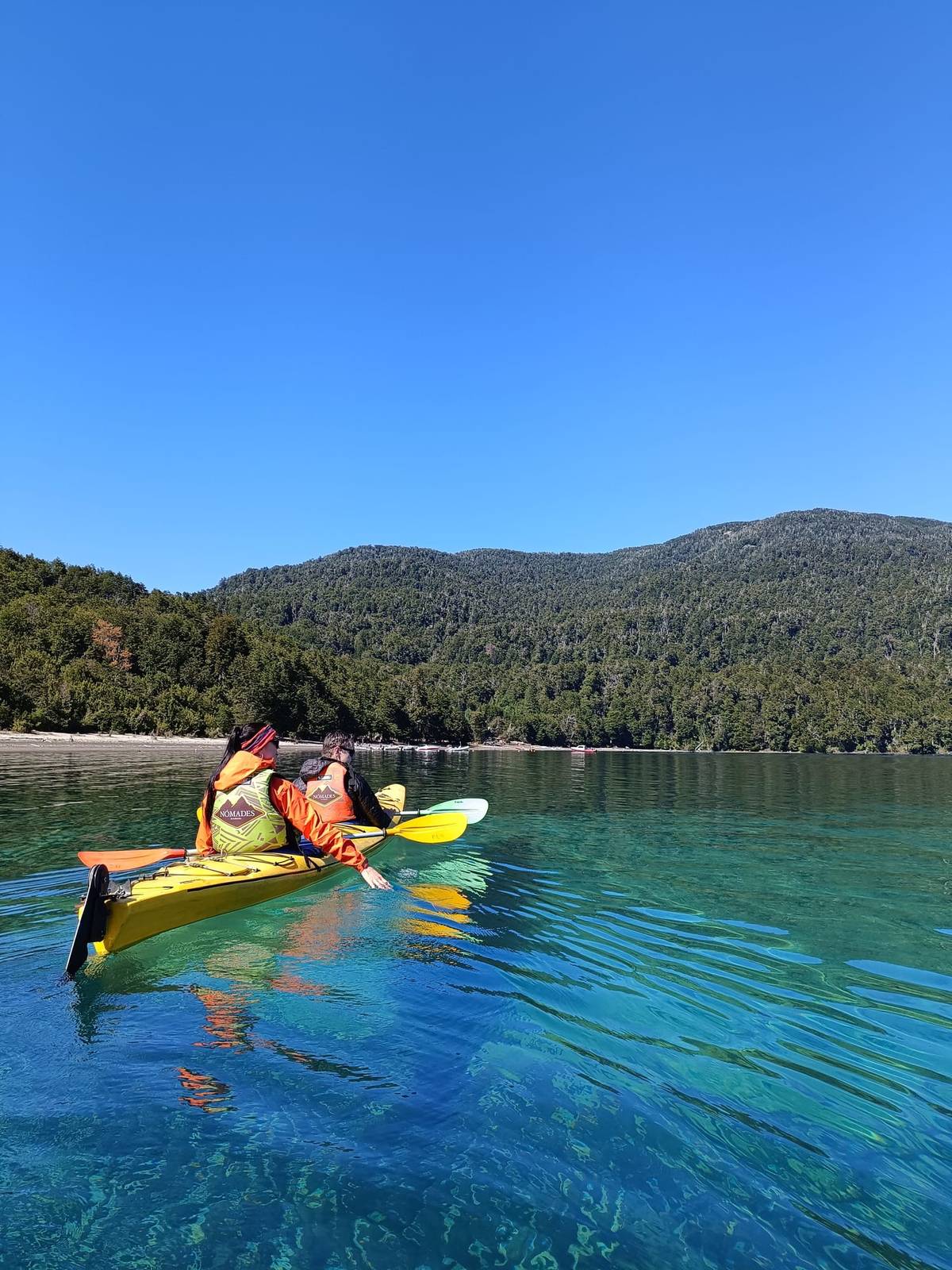 Dos personas en un kayak doble sobre agua turquesa y transparente, con la playa y el bosque de fondo