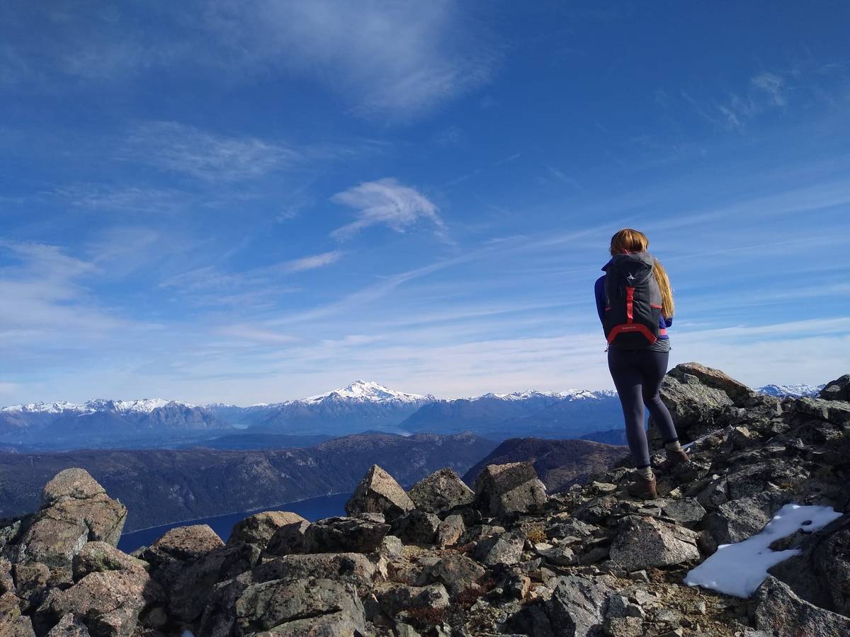 Persona con mochila parada sobre una cumbre rocosa, con un volcán nevado y la cordillera a lo lejos y un lago abajo