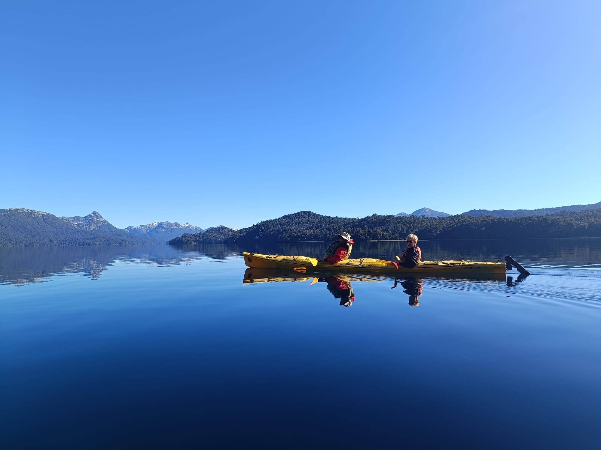 Dos personas en un kayak doble amarillo sobre un lago calmo como un espejo, con cerros cubiertos de bosque y picos nevados de fondo